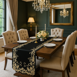 Elegant dining room with a dark wood table featuring a black and gold embroidered table runner, cream upholstered chairs, gold candleholders, white rose centerpiece, and a crystal chandelier reflecting in a large framed mirror.