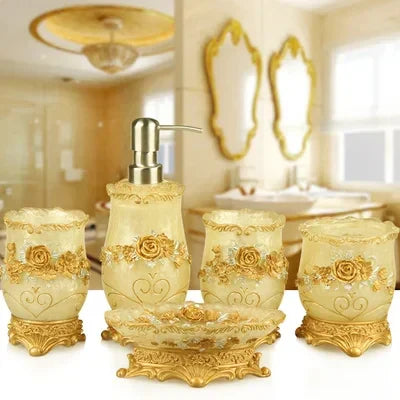 Ornate gold-tone ceramic bathroom accessories set—including soap dispenser, toothbrush holder, tumbler, cotton jar, and soap dish—with intricate rose and scroll detailing, displayed on a vanity in a luxurious cream-and-gold bathroom.