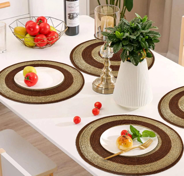 A modern dining table set with round brown and gold beaded placemats, featuring white plates, fresh vegetables, a potted plant centerpiece, a candle holder, and a fruit basket in a bright and elegant dining room.